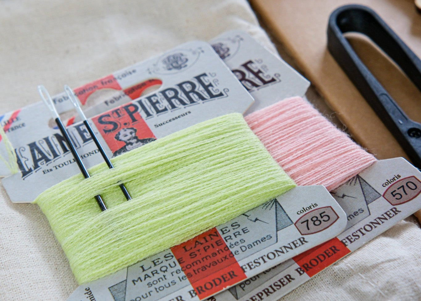 Two spools of embroidery floss on cards with brand names, placed on a white fabric background.