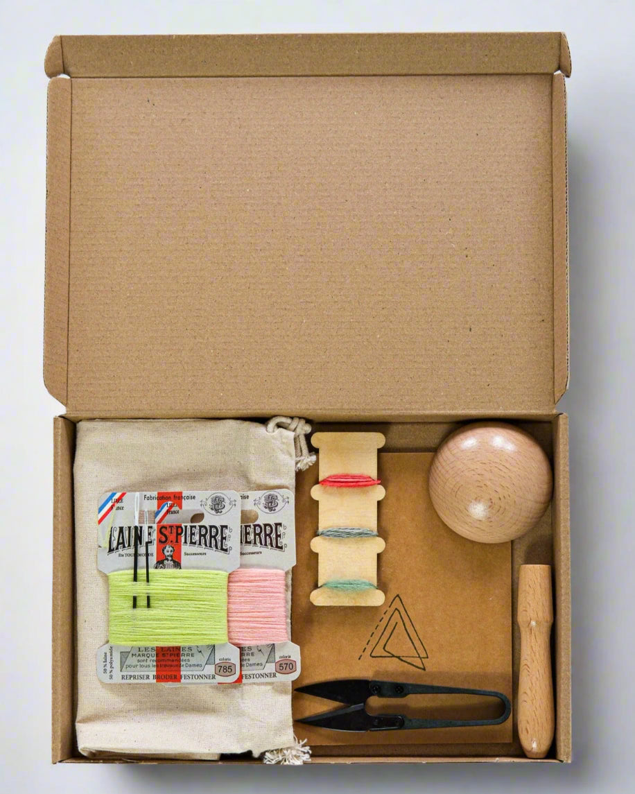Open cardboard box containing various mending items including a travel bag, an application brush, a darning mushroom, needles and other small objects on a white background.