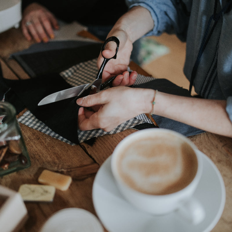 Person cutting a piece of fabric with scissors on a table with a cup of coffee and other items.