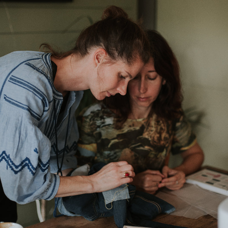 Two women sitting at a table with a box of items, sewing in a one on one class