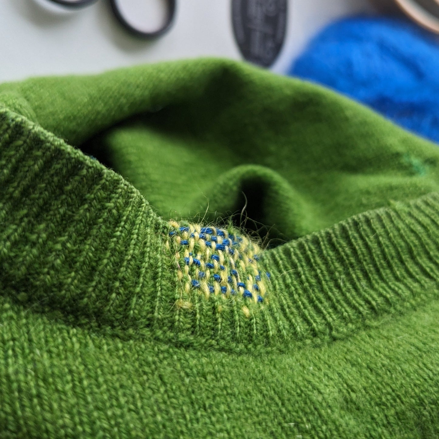 Close-up of a green knitted garment with a small embroidered visible mending design, surrounded by yarn and knitting tools on a white surface.