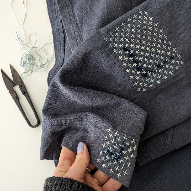 Dark denim jacket with cross stitch sashiko pattern embroidery held by a hand, scissors and thread in the background.