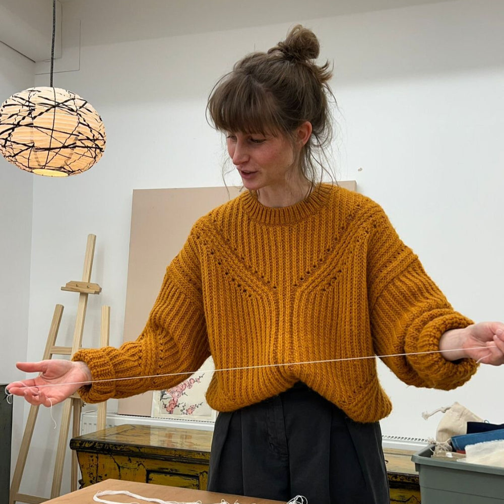 Woman in a mustard sweater working with yarn at a table in a room with art supplies.