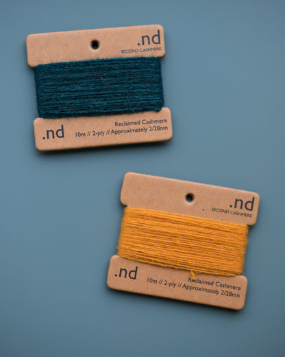 Two spools of cashmere thread, one green and one yellow, on cardboard labels with 'nd' branding against a blue background.