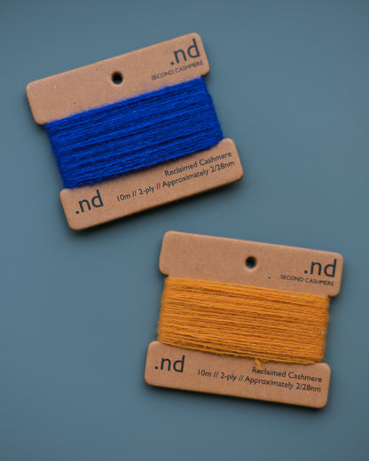Two spools of cashmere thread, one blue and one yellow, on brown cards with 'nd' branding against a gray background.