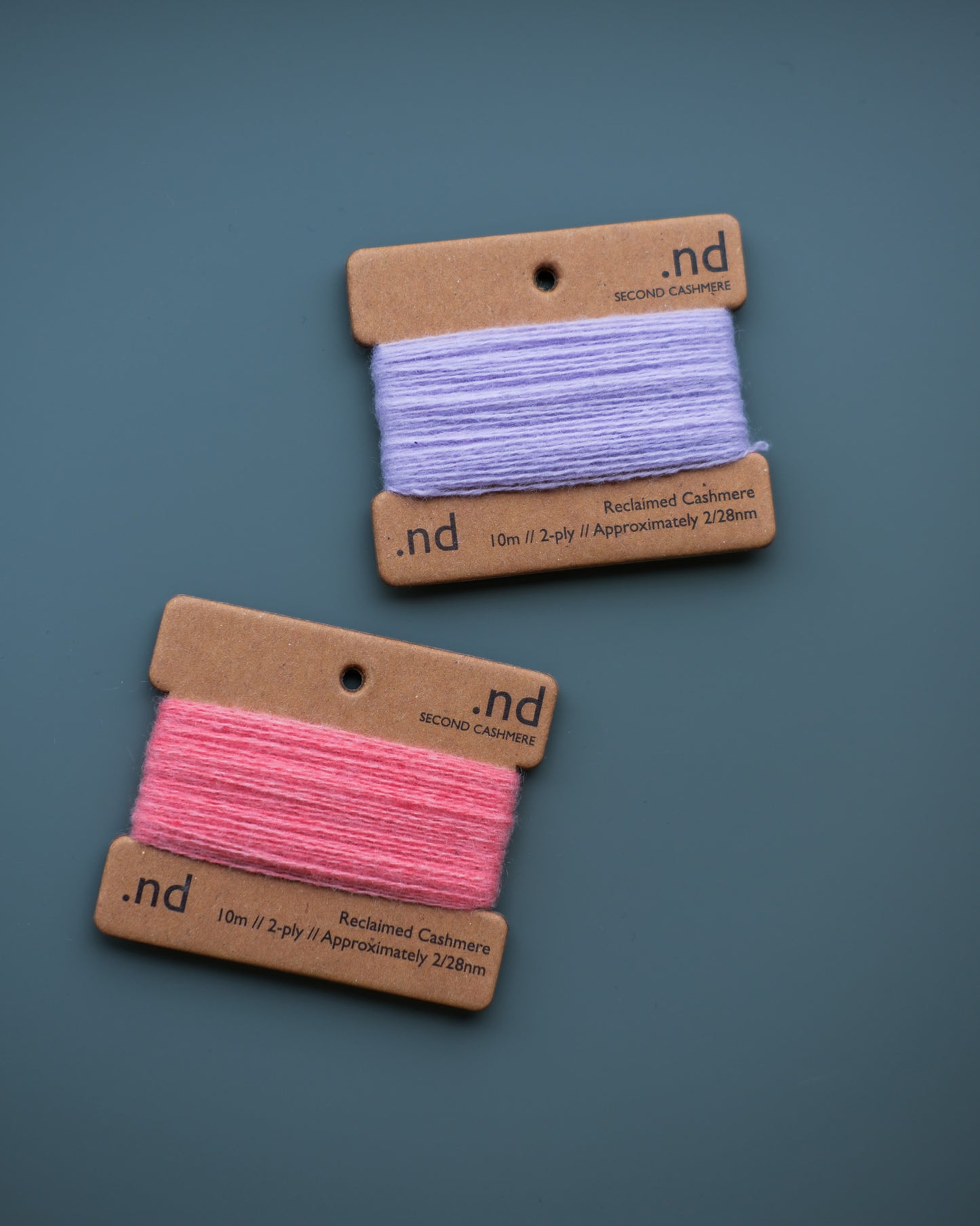 Two spools of cashmere thread, one pink and one purple, on brown cardboard labels with 'nd' branding against a blue background.