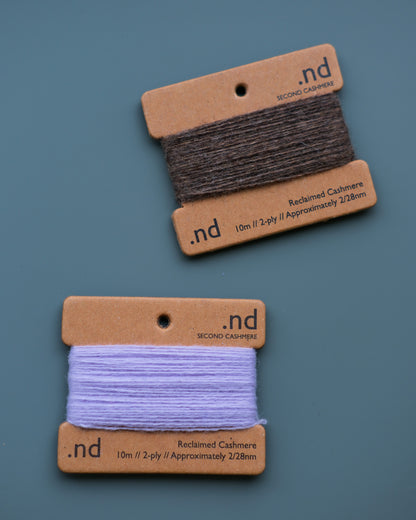 Two spools of reclaimed cashmere yarn on branded brown cards against a blue background