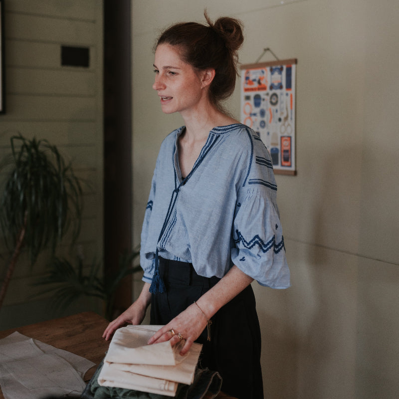 Woman holding a stack of fabric in a room with a plant and framed picture on the wall.