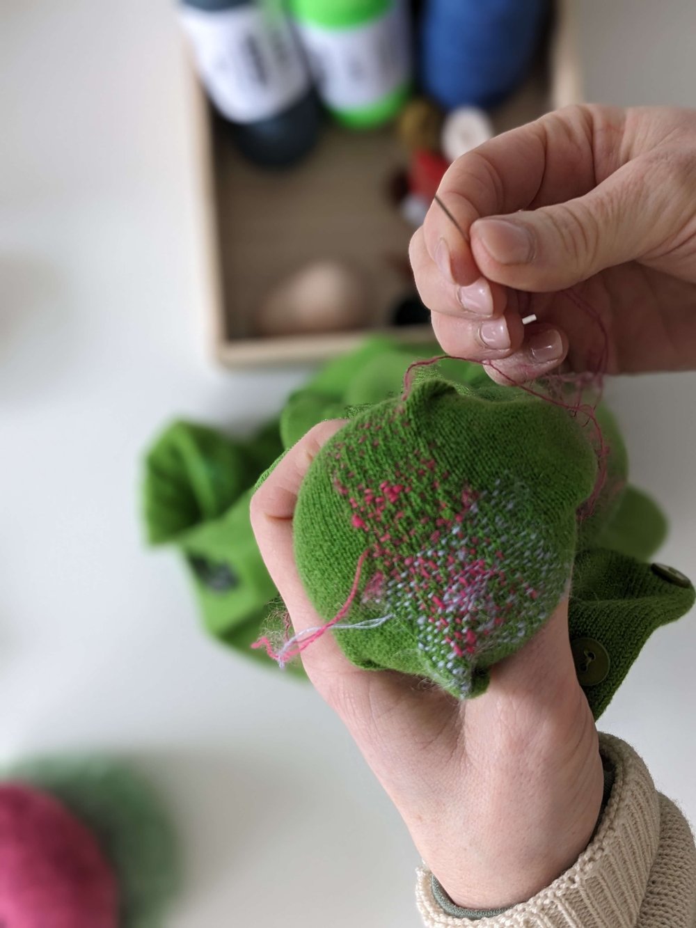 Hand holding a green textured object with pink threads, blurred background with skeins of thread.