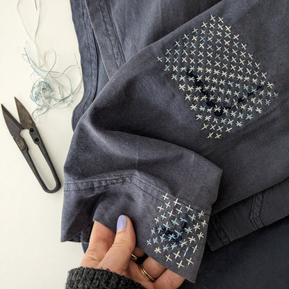 Dark denim jacket with cross stitch sashiko pattern embroidery held by a hand, scissors and thread in the background.